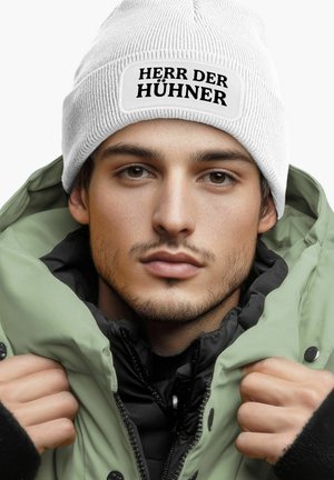White knit beanie with a black patch reading "HERR DER HÜHNERN." Worn with a green and black jacket, showcasing textured fabrics and layered styling.