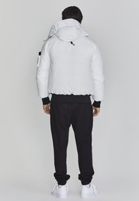 Hvid pufferjakke med hætte, der har elastiske mansjetter og et logo på kraven. Bæres over sorte sweatpants og hvide sneakers.