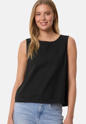 Kvinde iført ærmeløs sort top med blondekant og lyseblå jeans, smilende mod ensartet lysegrå baggrund.