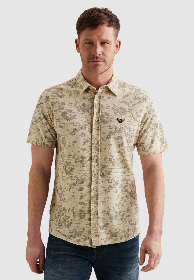 Homme portant une chemise à manches courtes beige à boutons avec un motif floral subtil et un petit logo de papillon sur la poitrine, associée à un jean foncé.