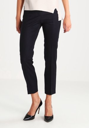 Frau trägt schlanke schwarze, knöchellange Hosen, kombiniert mit schwarzen, spitzen High-Heel-Schuhen und einem hellbeigen Oberteil mit Seitenschlitzen.