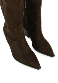 Botas de ante marrón hasta la rodilla con punteras puntiagudas y costuras decorativas. Cuentan con una cremallera lateral y un tacón cuadrado. Textura suave en general.