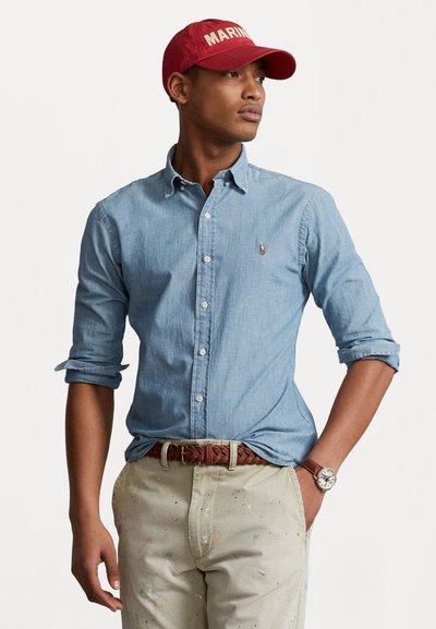 Lys blå chambray skjorte med knap-ned krave, rullede ærmer, cremefarvede bukser med malingspletter, brun fletbælte, rød "MARINE" kasket.
