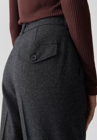 Vue arrière d'une personne portant un pantalon gris foncé avec des motifs et une poche arrière boutonnée, ainsi qu'un haut à manches longues côtelé marron.