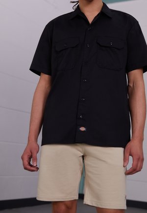 Personne portant une chemise noire à manches courtes boutonnée et un short beige, debout à l'intérieur contre un mur clair.
