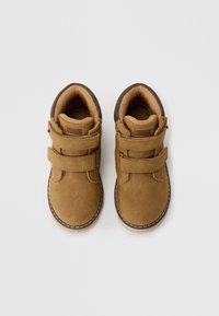 Zapatos de ante marrón con dos correas de Velcro, cuello acolchado y suela lisa. Presenta una etiqueta de logo en el lado y costuras claras contrastantes.