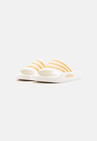 Λευκά σανδάλια Adidas τύπου slide με τρεις πορτοκαλί ρίγες και ανάγλυφο λογότυπο Adidas στο πλάι, εμφανίζονται σε ελαφρώς γωνιακή θέση πάνω σε λευκό φόντο.