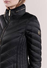 Giacca a vento nera con design a trapunta a zig zag, colletto alto e hardware dorato. Presenta un emblema sulla manica sinistra e tasche con zip.
