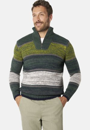 Man wearing a multicolored striped knit sweater with a zip-up collar and beige pants, hands in pockets, looking slightly to the side.