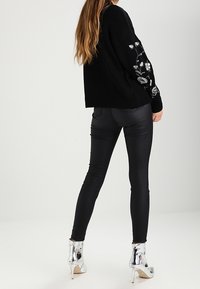 Femme debout, portant un pull noir orné de broderies florales, un pantalon en cuir noir moulant et des bottines argentées à talons hauts et brillantes.
