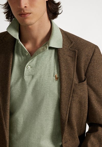 Rudas žirnelio rašto švarkas dėvimas ant šviesiai žalios polo marškinėlių. Polo turi apykaklę, tris mygtukus ir mažą siuvinėtą arkliuko logotipą.