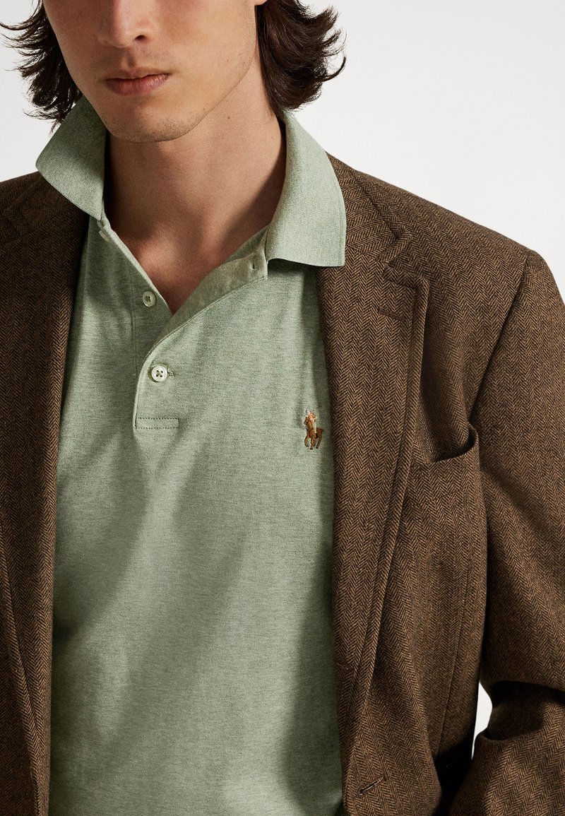 Brauner Fischgrät-Blazer über einem hellgrünen Poloshirt. Das Poloshirt hat einen Kragen, drei Knöpfe und ein kleines gesticktes Pferde-Logo.