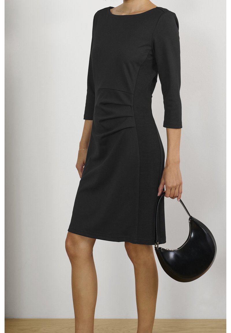 Femme en robe noire mi-longue aux genoux avec manches 3/4 tenant un sac à main noir en forme de croissant à ses côtés.