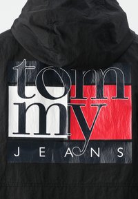 Zwarte jas met een groot gedrukt logo in vette typografie, gecombineerd met rood, wit en blauwe kleuren en glanzende accenten.