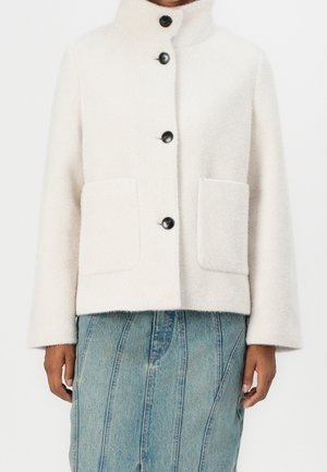 Manteau en laine blanche boutonné avec col montant et deux poches avant, porté sur une jupe en jean bleue surpiquée.