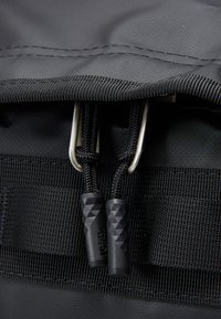 Svart ryggsekk med teksturert stoff, utstyrt med to sølvfargede metallklips, svarte paracord-glidelåser og gripeender med diamantmønster.