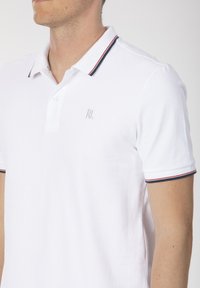 Polo shirt blanche en tissu texturé, avec un plastron à trois boutons et un col orné de rayures bleues et rouges. Petit logo brodé.