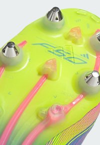 Bright yellow outsole of a soccer cleat, featuring metallic silver studs and pink accents. Stripes and logo are visible on the surface.