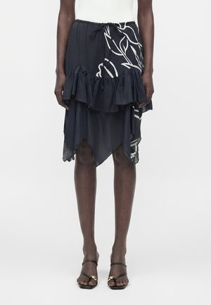 Czarna asymetryczna spódnica z białym kwiatowym wzorem i falbanką, noszona z czarnymi sandałami na otwartym nosku z ozdobami w postaci złotej klamry.