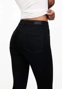 Osoba v černých skinny džínách a bílém bezrukávovém topu, zvedající tričko, aby odhalila oblast pasu.