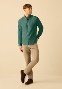 Cămașă lungă cu mânecă verde, cu nasturi, cu o croială slim, asociată cu pantaloni bej și cizme cu șireturi maro închis. Material neted, guler clasic.