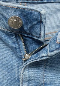 Giacca di denim con un lavaggio azzurro chiaro, dotata di un bottone a pressione argentato e di una cerniera in ottone. Dettagli di cuciture visibili con variazioni di texture.