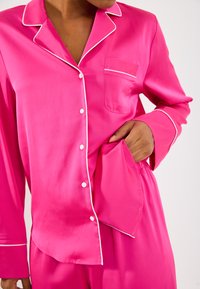 Ružový saténový pyžamový set s bielym lemovaním. Obsahuje košeľu na gombíky s vreckom a voľné nohavice s elastickým pásom.