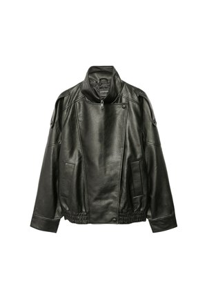 Femme portant une veste en cuir sombre oversize, un top court gris, des leggings capri noirs et des talons pointus noirs, debout devant un fond gris.
