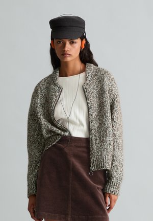 Junge Frau trägt eine graue Strickjacke, ein weißes Top, einen braunen Cordrock, eine schwarze Mütze und orange Kopfhörer vor einem schlichten Hintergrund.