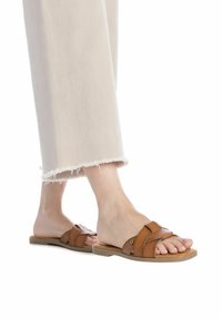 Sandalias deslizantes de cuero marrón con diseño de tiras trenzadas, suela plana y hardware mínimo. Combinadas con pantalones ligeros de dobladillo deshilachado.
