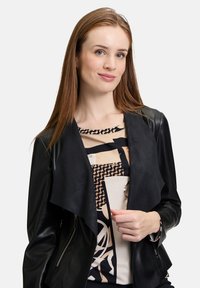 Veste en cuir synthétique noire avec un devant drapé et doux. Portée par-dessus un haut à motifs présentant des designs abstraits en beige et noir.