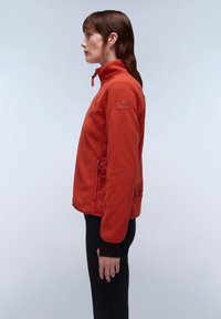 Giacca in fleece rossa con colletto alto, dotata di tasche laterali e dettagli con logo. Texture liscia e vestibilità rilassata, abbinata a pantaloni neri.