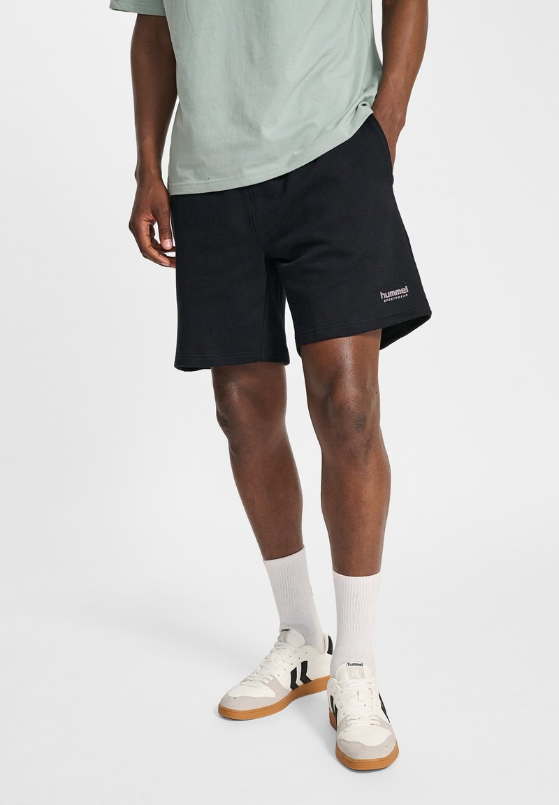 Shorts en coton noir à coupe décontractée, avec un logo brodé sur l'ourlet, associés à des chaussettes blanches et des baskets pour un look décontracté.