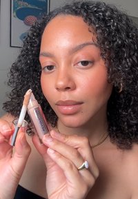 Un tube d'anticernes Revolution avec un applicateur transparent, montrant un liquide beige clair. Une femme tient le tube, affichant des cheveux ondulés et des bijoux.