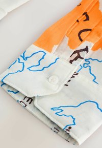 Manche de chemise repliée avec base blanche, motifs abstraits en lignes bleues, motifs orange et noirs, et un bouton blanc sur le poignet.