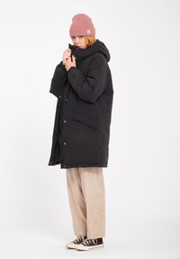 Zwarte pufferjas met een capuchon, voorzien van voorzakken, knoopsluiting en een gladde textuur. Gepaard met beige corduroybroeken en bruine sneakers.