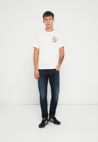 Camiseta de algodón blanca con estampado negro, combinada con jeans de mezclilla azul oscuro y zapatillas negras con rayas blancas. Look simple y casual.