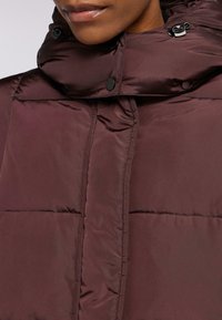 Giacca imbottita bordeaux con colletto alto, dotata di chiusura zip, bottoni a pressione e una texture liscia e lucente. Motivo di trapuntatura ben visibile.