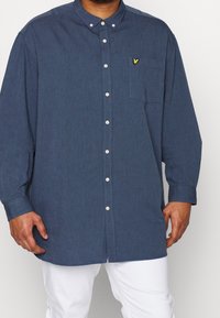 Niebieska koszula zapinana na guziki z długimi r�ękawami, kieszenią i małym żółtym haftowanym logo na piersi. Wykonana z tkaniny o fakturze.