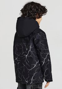 Giacca nera con cappuccio, caratterizzata da un motivo a fulmine bianco. Realizzata in tessuto liscio e resistente. Vestibilità casual e design di lunghezza media.