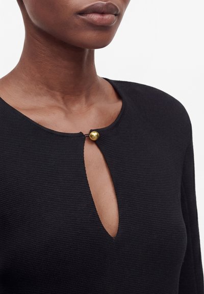 Gros plan d'une personne portant un haut noir texturé avec un col rond et un petit bouton doré fermant une ouverture en forme de trou de serrure.
