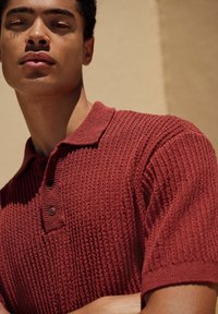 Polo de culoare roșie, texturat, cu guler, având o închidere cu trei butoane. Materialul pare a fi respirabil, cu un model tricotat.