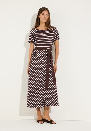 Femme portant une robe midi rayée marron et blanche à manches courtes, ceinture, et sandales noires à enfiler, debout contre un mur blanc.
