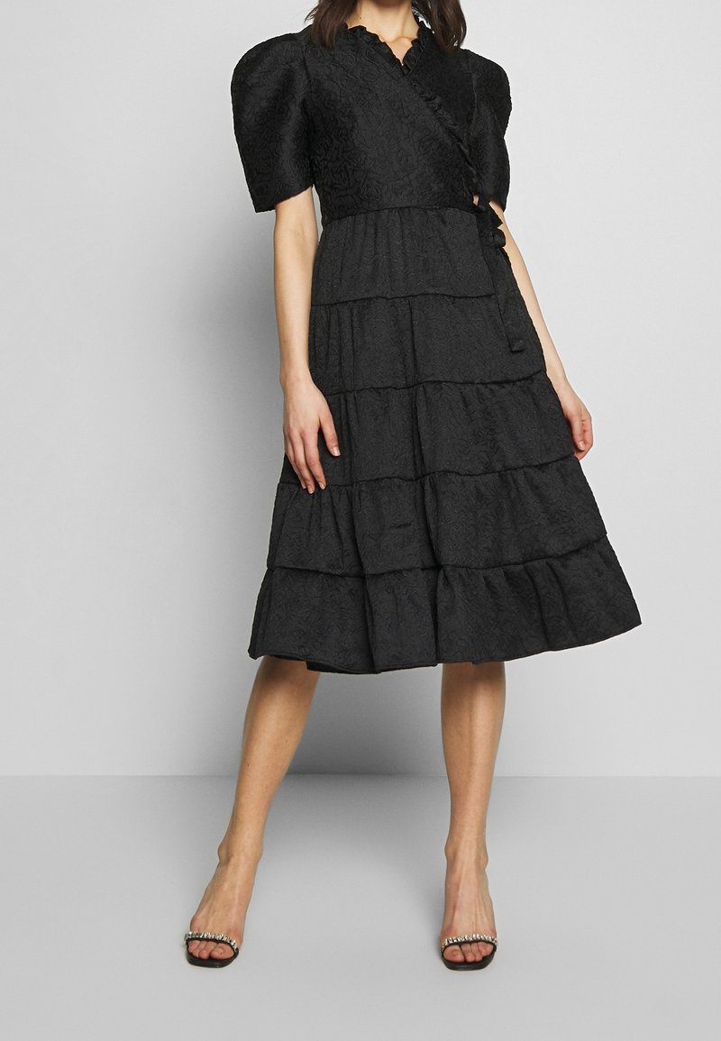 Robe noire à volants avec manches bouffantes et motif floral texturé. Comprend un design portefeuille et un détail d'encolure à volants.