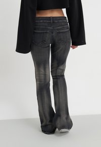 Persona vestida con jeans acampanados negros desgastados y un top negro de manga larga cortado, caminando sobre un suelo gris claro contra una pared blanca.