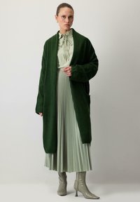 Long green wool cardigan over a light green blouse with a bow detail, paired with a pleated skirt and shiny green heeled boots.