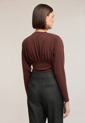 Kobieta o prostych brązowych włosach nosi bordową bluzkę z długim rękawem i marszczonym tyłem oraz wysokiej talii ciemnoszare spodnie, stoi bokiem na jednolitym tle.