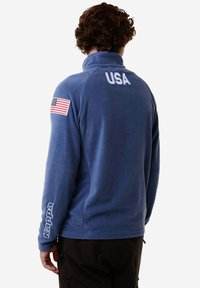 Giacca in pile blu con colletto alto, "USA" stampato sul retro e un patch della bandiera americana sulla manica sinistra. Logo sulla manica destra.