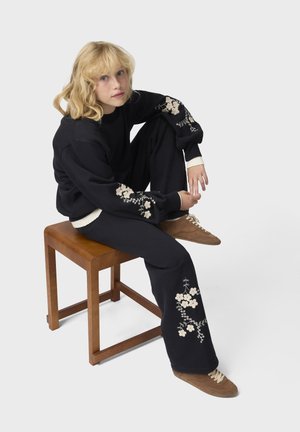 Femme blonde en sweat-shirt noir brodé de fleurs et pantalon, assise sur un tabouret en bois avec un genou relevé, portant des chaussures marron à lacets.