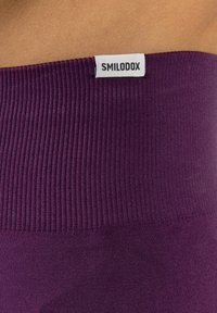 Cinturilla morada acanalada con una textura suave, que presenta una etiqueta blanca grabada con "SMILODOX" en el borde superior.